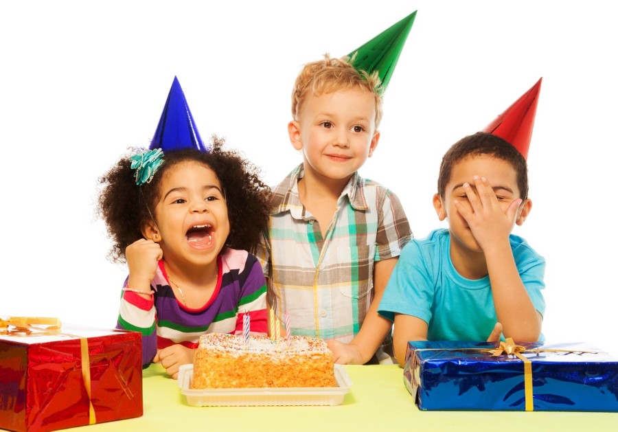 3 kinderen met feesthoedjes op staan aan een tafel met een verjaardagstaart en 2 ingepakte cadeautjes voor kinderen (een blauw en een rood)